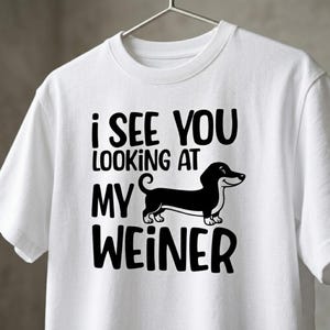 May include: A white short-sleeved t-shirt featuring a black graphic of a dachshund dog. The text printed in black above and below the dog reads, I SEE YOU LOOKING AT MY WEINER. The shirt is displayed on a metal hanger against a neutral background.