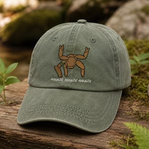 May include: A sage green baseball cap featuring an embroidered tan-colored spider-like creature with a small green striped band on one leg. Below the creature, the text "AMAZE AMAZE AMAZE" is embroidered in white thread on the front panel.