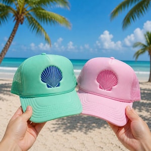 May include: A mint green trucker hat with a dark blue seashell graphic and a light pink trucker hat with a matching pink seashell graphic. Both hats feature a structured front panel, a curved brim, and a breathable mesh back for ventilation.