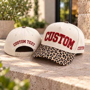 May include: Two off-white baseball caps with leopard print brims and top buttons. The front cap features the word CUSTOM in red embroidered letters, while the back cap displays the text CUSTOM TEXT in red. Both caps have adjustable snapback closures.