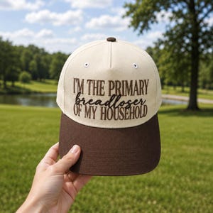 May include: A cream-colored baseball cap with a dark brown brim and a dark brown embroidered text that reads I'M THE PRIMARY breadloser OF MY HOUSEHOLD. The cap features a button on top and white eyelets for ventilation on the front panels.