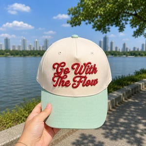 May include: A baseball cap featuring a cream-colored crown and a light green brim. The front of the cap displays the text Go With The Flow embroidered in a bold, raised red script font. The cap includes a small green button at the top center.