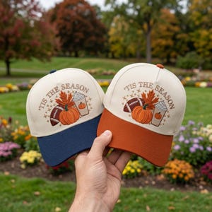 May include: Two baseball caps with off-white crowns and colored brims, one navy blue and one burnt orange. Each features a graphic with a football, a pumpkin, a coffee cup, and autumn leaves, with the text 'TIS THE SEASON' printed in brown.