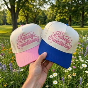 May include: Two five-panel trucker hats with off-white crowns and pink or blue brims. Each hat features the embroidered pink text "Barely Functioning" in a cursive font, accented by small star shapes, with a matching button on the top crown.