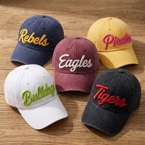 May include: Five baseball caps in various colors including navy blue with yellow Rebels text, maroon with white Eagles text, yellow with pink Pirates text, white with green Bulldogs text, and black with red Tigers text, all featuring embroidered lettering.