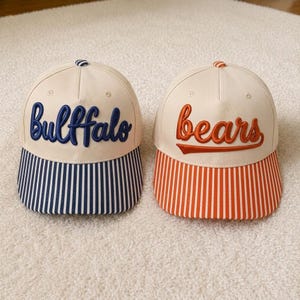 May include: Two baseball caps with off-white crowns. The left cap features the word "Bullfalo" in blue embroidery and a blue-and-white striped brim. The right cap features the word "bears" in orange embroidery and an orange-and-white striped brim.