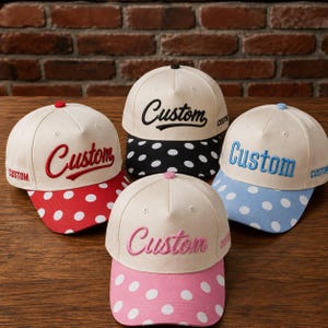 May include: Four baseball caps with cream-colored crowns and brims featuring white polka dots. Each cap displays the word Custom in embroidered script on the front and side. The brims and embroidery are red, black, light blue, and pink respectively.