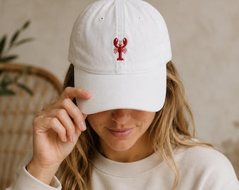 May include: A white baseball cap featuring a small, centered red lobster embroidery on the front panel. The cap has a structured crown, a curved brim, and a classic six-panel design with a button at the top, providing a clean and simple aesthetic.