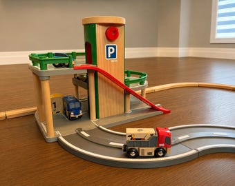 May include: A wooden toy parking garage with a central tower featuring a red knob and a blue P sign. The structure includes green railings, a red ramp, and multiple levels. A blue truck and a red truck are positioned on the gray plastic road tracks.