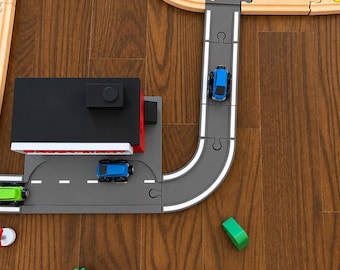 May include: A wooden toy train track set featuring gray road segments, a black and red building, and small blue and green toy cars. The set includes curved and straight wooden tracks with interlocking puzzle-style connectors.