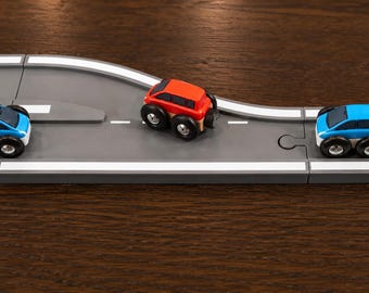 May include: A toy set featuring three small cars, two blue and one red, positioned on a gray modular road track. Each car has four black wheels and a compact body design. The road segments are connected with puzzle-like joints and feature white lane markings.