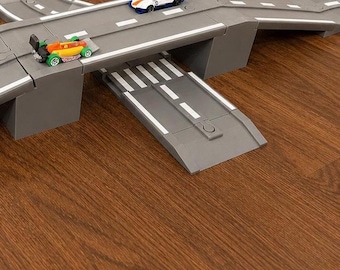 May include: A gray plastic toy car track system featuring elevated road sections, a ramp, and white painted lane markings. Two small die-cast toy race cars, one orange and green and one white and blue, are positioned on the track surface.