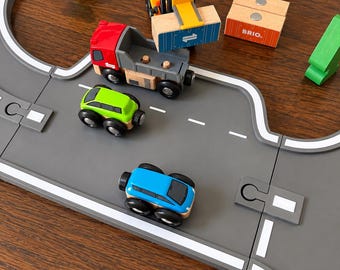 May include: A wooden toy set featuring a gray road track, a red dump truck, a yellow forklift, a green car, a blue car, and an orange cargo block labeled BRIO. The set includes green wooden trees and a blue cargo block with the number one.