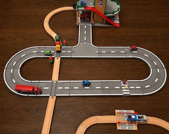 May include: A toy vehicle playset featuring a gray modular road system with white lane markings, connected to wooden train tracks. The set includes a multi-level parking garage, a red truck, several small cars, and miniature figures.