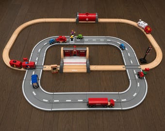 May include: A BRIO wooden train track set featuring a gray plastic road loop, a central station building, a red tunnel, and various small toy vehicles including cars, a truck, and a train engine with a carriage, all arranged on a flat surface.