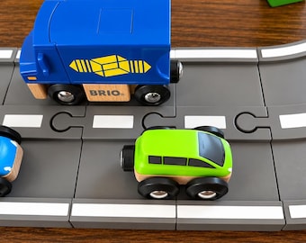 May include: A blue wooden toy truck with a yellow geometric logo and the word BRIO printed on the side, positioned on a gray plastic road track next to a small lime green wooden toy car with black wheels and a black windshield.
