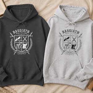 Puede incluir: Una sudadera con capucha gris carbón oscuro y una sudadera gris jaspeado claro, ambas con un estampado gráfico en blanco y negro de un escudo con el texto Basgiath War College Navarre y BWC, incluyendo iconos de un dragón y espadas.