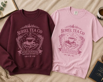 May include: A maroon long-sleeve sweatshirt and a light pink short-sleeve t-shirt, both featuring a graphic print of hands holding a stack of books and a teacup, with the text "SURIEL TEA CO" and "HOTTEST TEA IN PRYTHIAN AND EVEN HOTTER FAE".