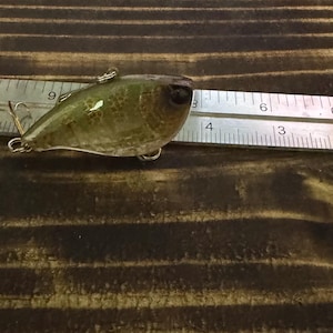 May include: A green and brown speckled fishing lure with a dark eye and two metal hooks, positioned next to a metal ruler that shows the lure is approximately 2 inches in length. The lure has a smooth, glossy finish and a rounded body shape.