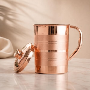 May include: A cylindrical copper pitcher with a polished finish and a matching lid placed to its left. The pitcher features a curved handle and horizontal bands of brushed texture alternating with smooth, reflective copper surfaces.