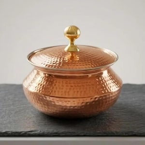 May include: A round, hammered copper serving bowl with a matching lid. The bowl features a wide, curved body and a polished, metallic finish. A small, spherical brass-colored knob sits atop the center of the lid, providing a contrast in color.