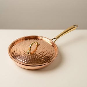 May include: A round copper frying pan with a hammered texture on the lid and a polished gold-tone metal handle. The lid features a matching gold-tone handle centered on top. The pan has a smooth, reflective copper finish and a sturdy construction.