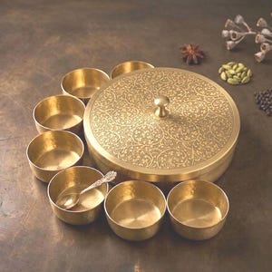 Könnte beinhalten: Eine runde Gewürzdose aus Messing mit einem floral gravierten Deckel und einem zentralen Knauf, umgeben von sieben kleinen passenden Messingschalen. Eine Schale enthält einen kleinen Messinglöffel. Das Set ist kreisförmig angeordnet.