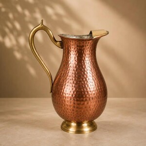 May include: A copper pitcher with a hammered texture, featuring a curved brass handle and a brass base. The pitcher has a wide, rounded body that tapers towards the top, ending in a flared spout with a smooth brass rim detail.