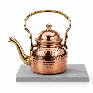 May include: A hammered copper teapot featuring a polished gold-tone curved spout, a matching gold-tone arched handle, and a domed lid with a small knob. The teapot has a rounded body with a textured surface and sits on a flat grey stone base.