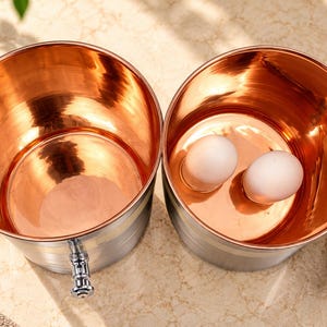 May include: Two cylindrical containers with polished copper interiors and brushed silver exteriors. One container is empty, while the other holds two white eggs and has a matching silver lid resting beside it. Each vessel features a small silver handle.