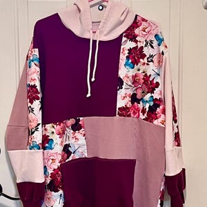 May include: A long-sleeve hooded sweatshirt featuring a patchwork design of solid deep plum, light pink, and a floral print fabric with pink flowers and blue butterflies. The hoodie includes a front kangaroo pocket, a drawstring, and ribbed cuffs.