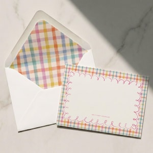 Op de afbeelding: Een witte envelop met een kleurrijke geruite voering, naast een bijpassende kaart. De kaart heeft een geschulpte roze rand en een lege ruimte voor tekst. Het ruitpatroon bevat tinten roze, geel, blauw en groen.