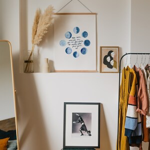 Op de afbeelding: Een wit ingelijst print met een aquarel-illustratie van de maan in verschillende fasen. De tekst luidt "De maan herinnert me eraan dat je nog steeds heel bent je bent groter dan je fasen je bent licht". De print hangt aan een witte muur met een gedroogd pampasgras in een glazen vaas.