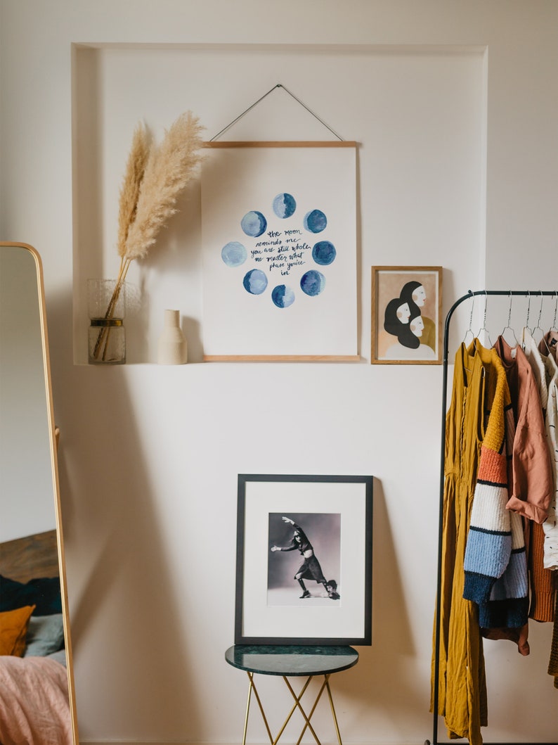 Op de afbeelding: Een wit ingelijst print met een aquarel-illustratie van de maan in verschillende fasen. De tekst luidt "De maan herinnert me eraan dat je nog steeds heel bent je bent groter dan je fasen je bent licht". De print hangt aan een witte muur met een gedroogd pampasgras in een glazen vaas.