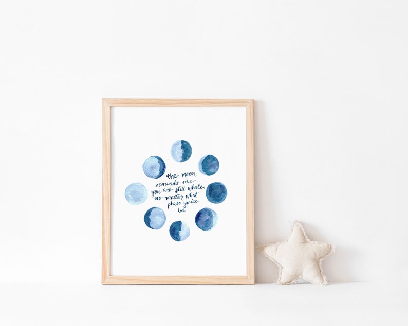 Op de afbeelding: Een ingelijst print met een aquarel-illustratie van verschillende maanfasen in tinten blauw. De tekst "the moon reminds me you are still whole, no matter what phase you're in" is geschreven in zwarte inkt.