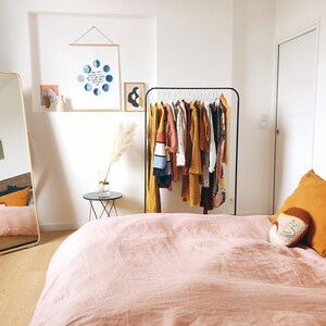 Op de afbeelding: Een slaapkamer met een roze linnen dekbedovertrek op een bed met een houten hoofdeinde. Er is een kledingrek met een verscheidenheid aan kleding die eraan hangt, een spiegel in de volle lengte en een klein bijzettafeltje met een vaas met gedroogd pampasgras.