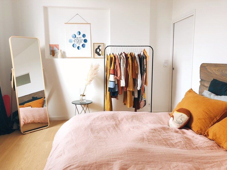 Op de afbeelding: Een slaapkamer met een roze linnen dekbedovertrek op een bed met een houten hoofdeinde. Er is een kledingrek met een verscheidenheid aan kleding die eraan hangt, een spiegel in de volle lengte en een klein bijzettafeltje met een vaas met gedroogd pampasgras.