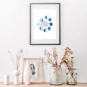 Op de afbeelding: Een ingelijst print met een aquarel-illustratie van de maan in verschillende fasen. De tekst luidt: "De maan herinnert ons eraan dat zelfs in de duisternis, er schoonheid en licht is dat we niet altijd kunnen zien, maar altijd kunnen voelen."