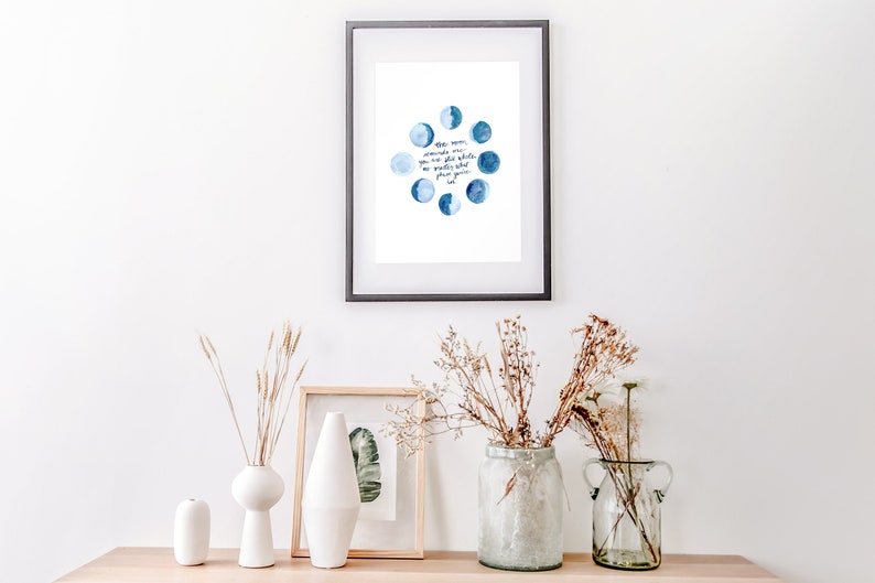 Op de afbeelding: Een ingelijst print met een aquarel-illustratie van de maan in verschillende fasen. De tekst luidt: "De maan herinnert ons eraan dat zelfs in de duisternis, er schoonheid en licht is dat we niet altijd kunnen zien, maar altijd kunnen voelen."