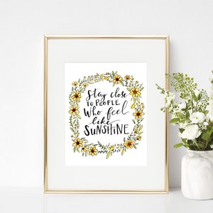 Könnte beinhalten: Ein gerahmter Druck mit weißem Hintergrund und einem Kranz aus gelben Blumen, der den Text "Bleib Menschen nahe, die sich wie Sonnenschein anfühlen" umgibt.