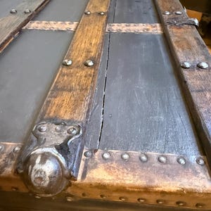 Puede incluir: Primer plano de un ba&uacute;l antiguo con aspecto envejecido. El ba&uacute;l presenta paneles grises oscuros enmarcados por vigas de madera envejecida y detalles met&aacute;licos. Remaches y esquinas met&aacute;licas decorativas a&ntilde;aden a la est&eacute;tica vintage.