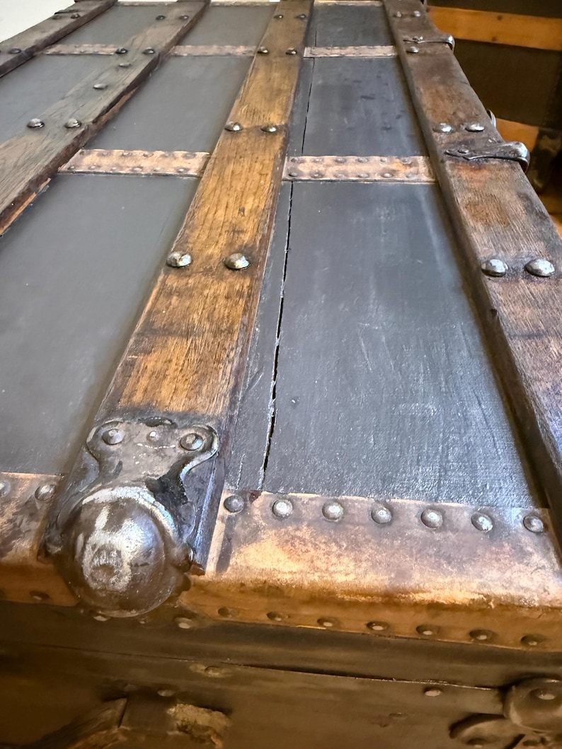 Puede incluir: Primer plano de un ba&uacute;l antiguo con aspecto envejecido. El ba&uacute;l presenta paneles grises oscuros enmarcados por vigas de madera envejecida y detalles met&aacute;licos. Remaches y esquinas met&aacute;licas decorativas a&ntilde;aden a la est&eacute;tica vintage.