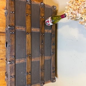 Puede incluir: Un ba&uacute;l de madera antiguo con detalles met&aacute;licos y una superficie gris oscuro. El ba&uacute;l tiene listones de madera verticales y protectores de esquina met&aacute;licos. Una botella de vino con etiqueta roja est&aacute; colocada.