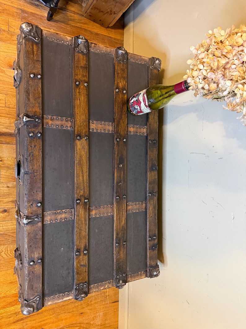 Puede incluir: Un ba&uacute;l de madera antiguo con detalles met&aacute;licos y una superficie gris oscuro. El ba&uacute;l tiene listones de madera verticales y protectores de esquina met&aacute;licos. Una botella de vino con etiqueta roja est&aacute; colocada.