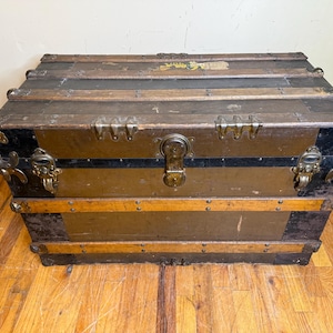 May include: An antique trunk with a weathered appearance. The trunk features a rectangular shape with metal hardware, including corner protectors and a central latch. The exterior is a mix of brown, black, and gold tones, with wooden slats and metal accents.