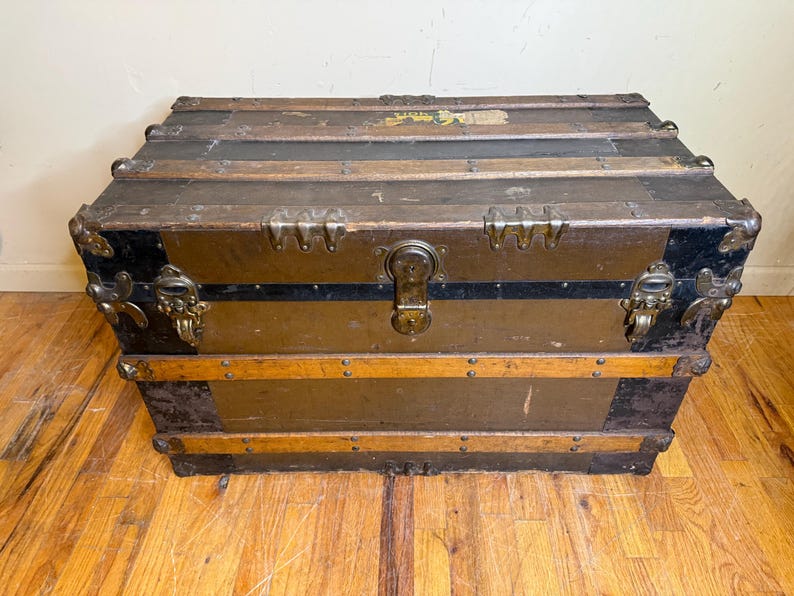 May include: An antique trunk with a weathered appearance. The trunk features a rectangular shape with metal hardware, including corner protectors and a central latch. The exterior is a mix of brown, black, and gold tones, with wooden slats and metal accents.