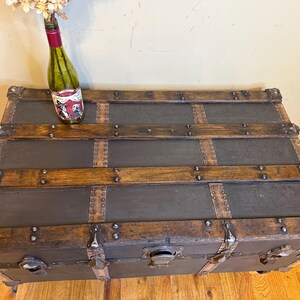 Puede incluir: Un ba&uacute;l antiguo con detalles en metal oscuro y madera. El ba&uacute;l tiene protectores de esquina de metal y un pestillo. Una botella de vino con una etiqueta decorativa y flores secas encima.