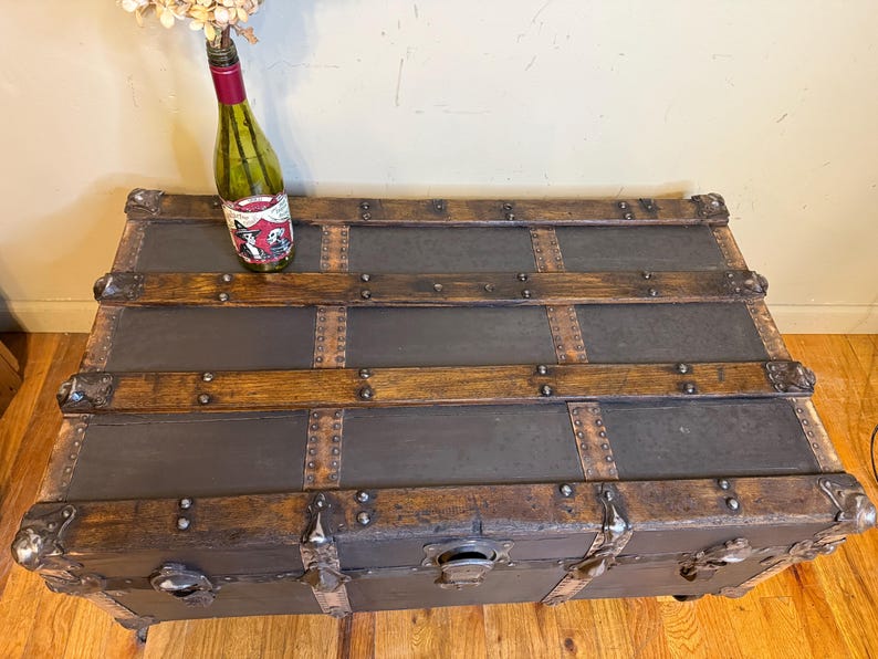 Puede incluir: Un ba&uacute;l antiguo con detalles en metal oscuro y madera. El ba&uacute;l tiene protectores de esquina de metal y un pestillo. Una botella de vino con una etiqueta decorativa y flores secas encima.