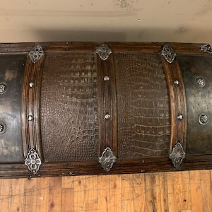 Pode incluir: Um ba&uacute; de madeira vintage com uma tampa de couro marrom com relevo de crocodilo. O ba&uacute; tem detalhes em metal e tachas decorativas de metal.