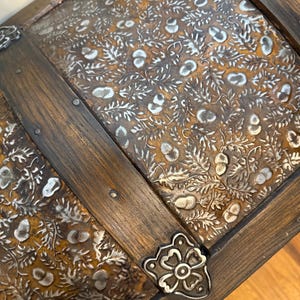 May include: Close-up of a vintage wooden chest with ornate metal detailing. The chest features a dark brown wood frame and a decorative metal panel with a floral pattern. The metal accents are silver-colored, and the chest has metal corner protectors.